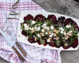 Pyszne carpaccio z buraka z fetą, rukolą i słonecznikiem na białym talerzu, obok sztućce i kraciasty ręcznik.
