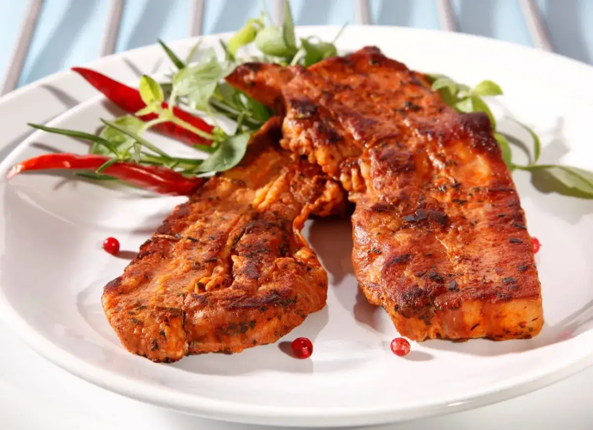 Dwa soczyste kawałki boczku na grilla, przyprawione i podane z papryczkami chili i zielonymi ziołami.
