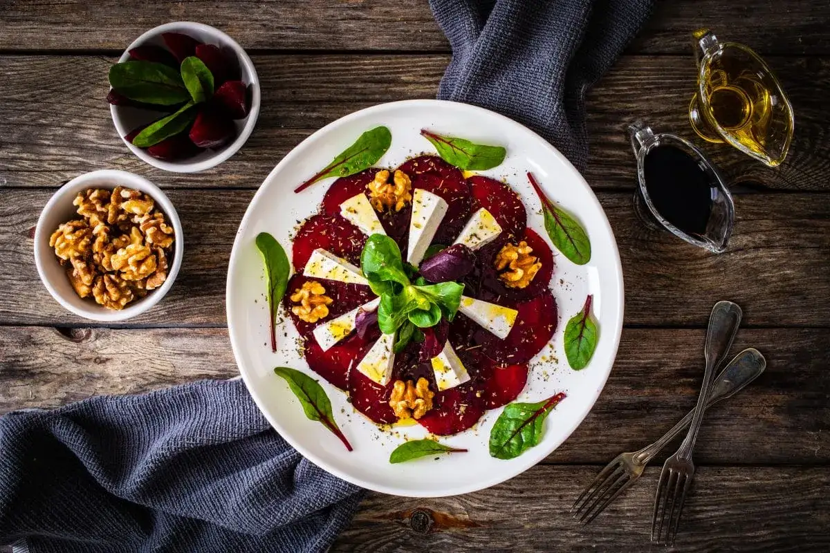 Eleganckie carpaccio z buraka z serem feta, orzechami włoskimi i świeżymi ziołami, podane na białym talerzu.