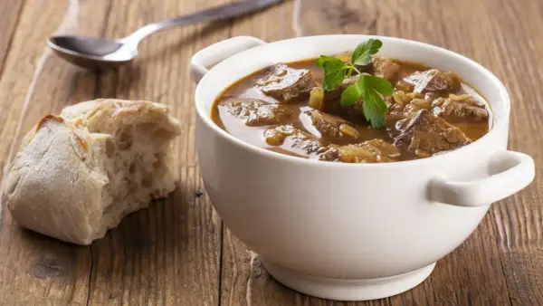 Aromaticzny gulasz wołowy w białej miseczce, ozdobiony natką pietruszki, obok kromka chleba i łyżka.